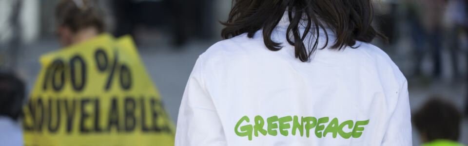 Une militante de Greenpeace de dos portant un habit Greenpeace. Au second plan un bannière sur laquelle on distingue 