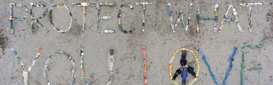 La photo est prise du ciel. Les militants ont écrit en très grand sur le sable les mots “Protect What You Love” (Protège ce que tu aimes) avec des déchets trouvés sur la plage. Ils ont formé un signe de peace and love ave le O du mot Love dont leurs corps forment les barres intérieures. - Opération nettoyage de déchets sur une plage de l'Arctique (Svalbard).
