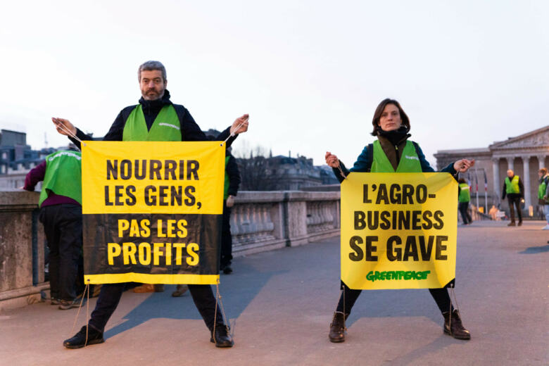 Nourrir les gens, pas les profits / L’agro-industrie se gave : deux messages sur des banderoles tenues par le directeur et une militante de Greenpeace en soutien aux agriculteurs, face à la crise agricole