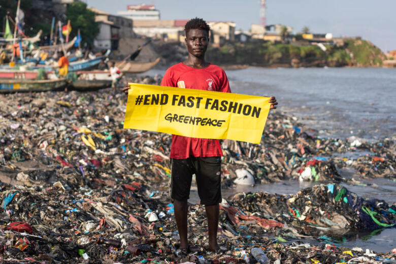 Un jeune homme tient une banderole "stop fast fashion" sur une plage recouverte de déchets textiles au Ghana.